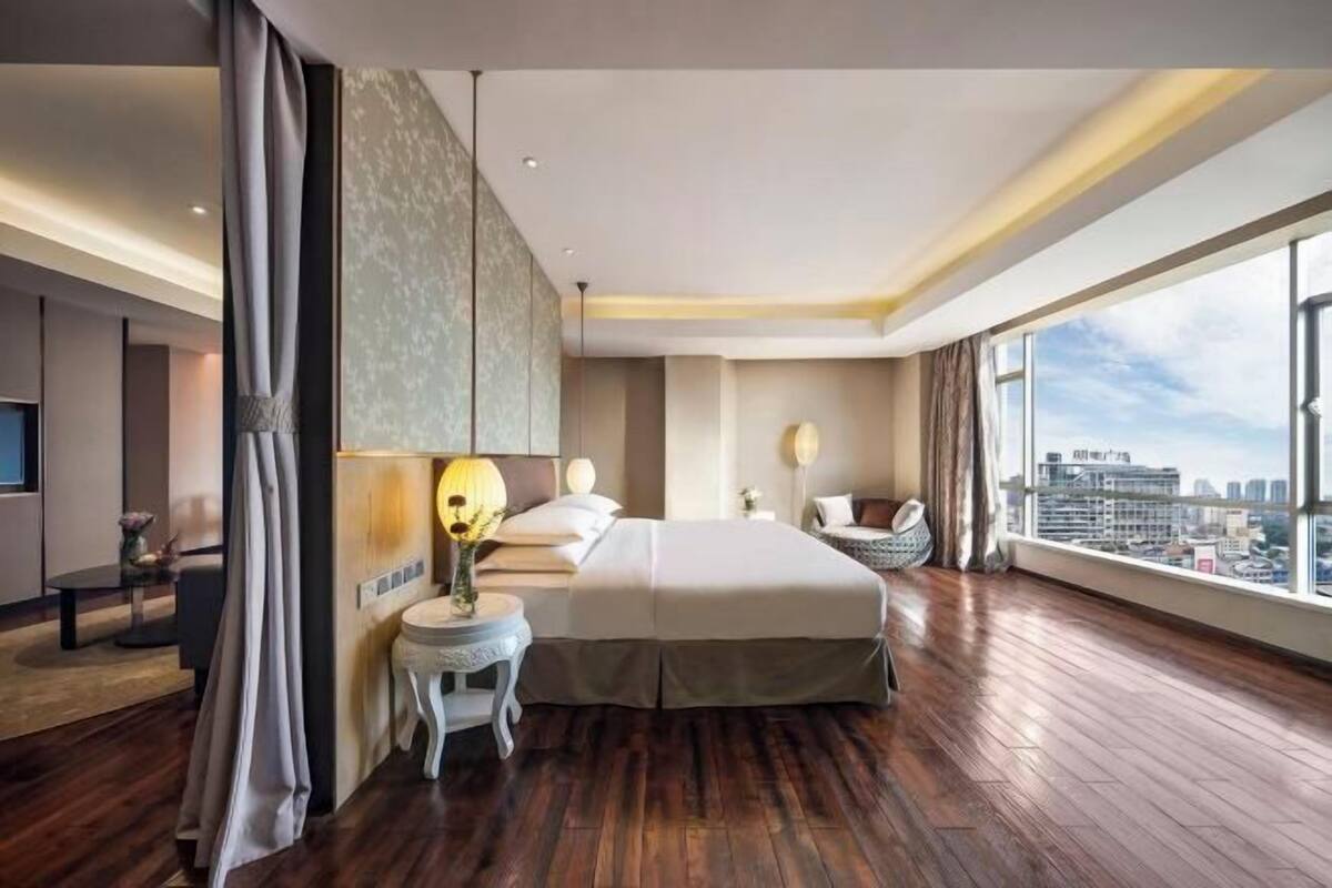 Een moderne hotelkamer met een groot bed, een klein bijzettafeltje, een nachtkastje, een televisie en een groot raam met uitzicht over de stad.