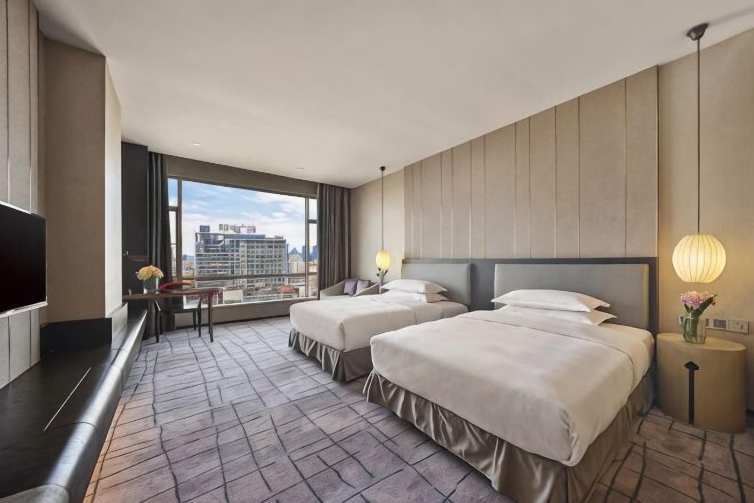 Habitación de hotel con dos camas, televisión, escritorio y ventana grande con vista a la ciudad.