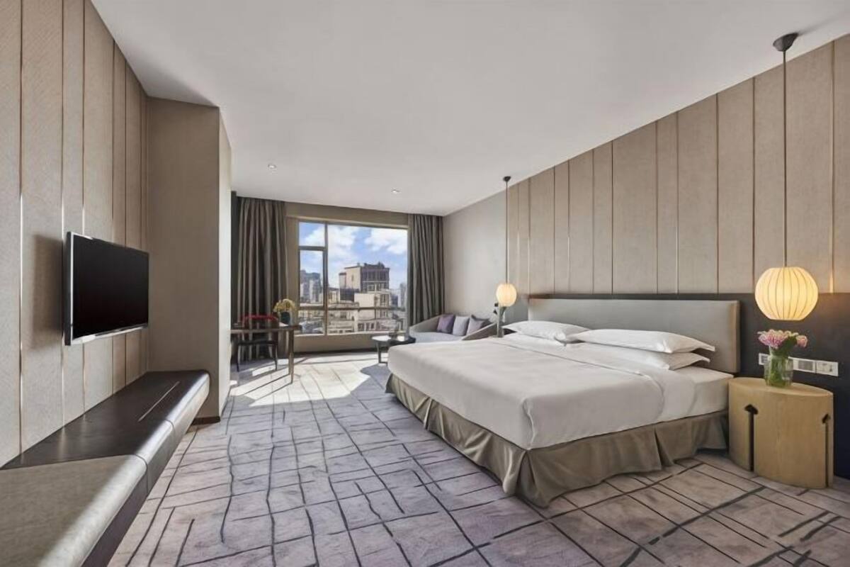Een hotelkamer met een groot bed, een flatscreen televisie, een zithoek met een tafel, en uitzicht op de stad.