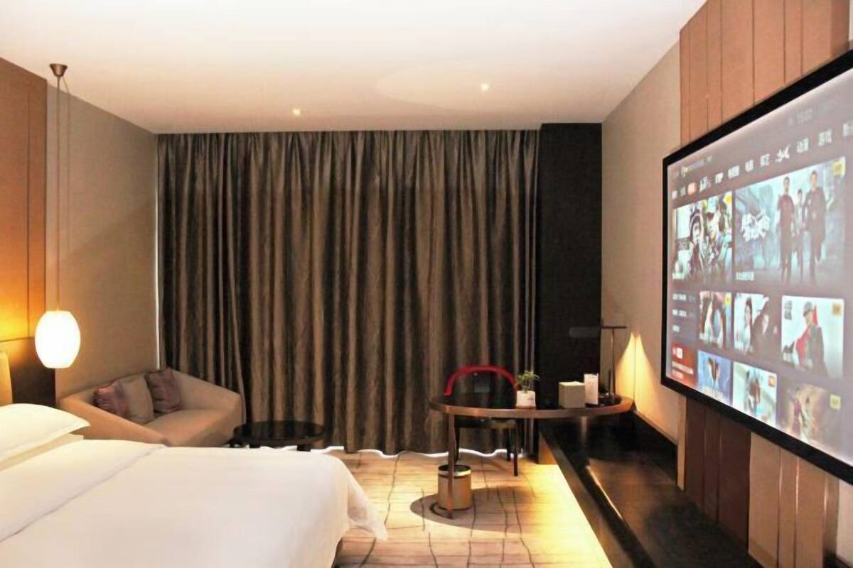 Een hotelkamer met een bed, een zithoek met een tafel en stoelen, een groot scherm waarop meerdere afbeeldingen worden getoond, en een gordijn.