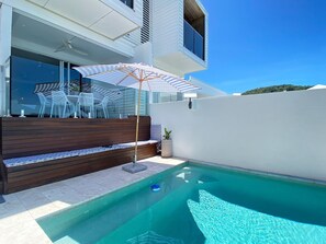 Un espace piscine extérieur moderne, doté d’une terrasse en bois, d’un coin salon avec des chaises et d’un grand parasol.