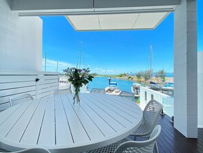 A white wooden deck with a round table and chairs, overlooking a marina with boats.