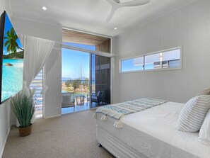 A bedroom with a large bed, a potted plant, a view of the ocean, and a balcony with a chair.