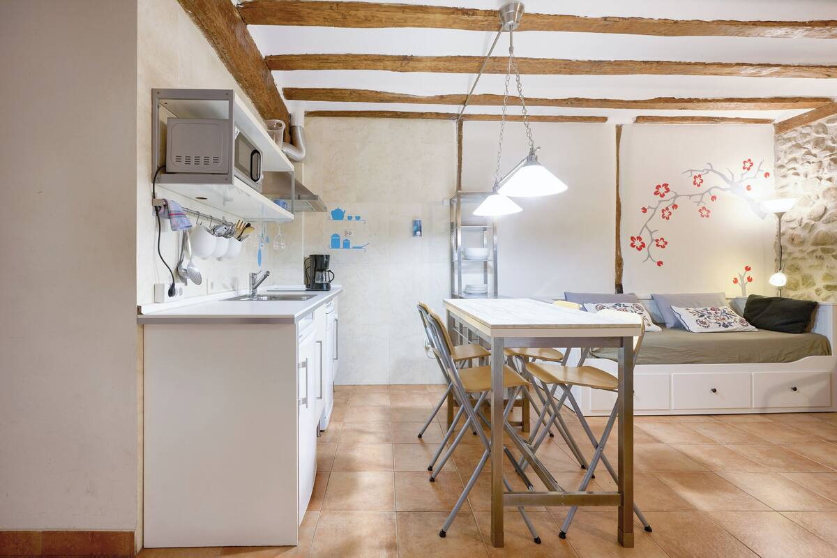 Une cuisine compacte avec des meubles blancs, une table à manger avec des chaises et un lit agrémenté d’un mur décoratif.
