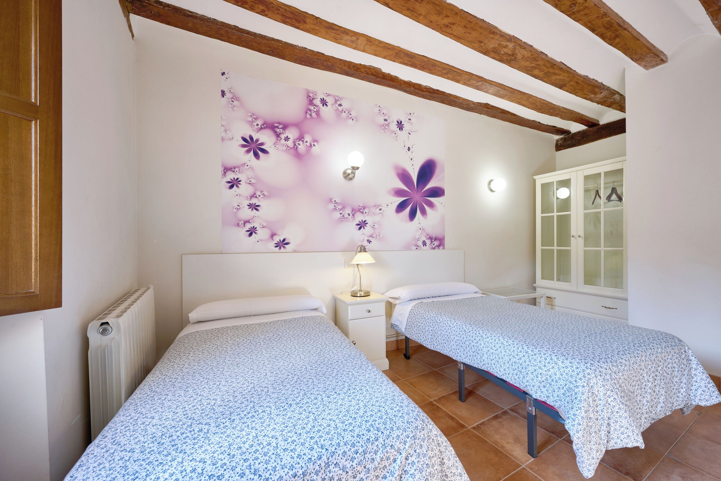 A bedroom with two beds, a nightstand, a wall-mounted lamp, and a cabinet with a window.