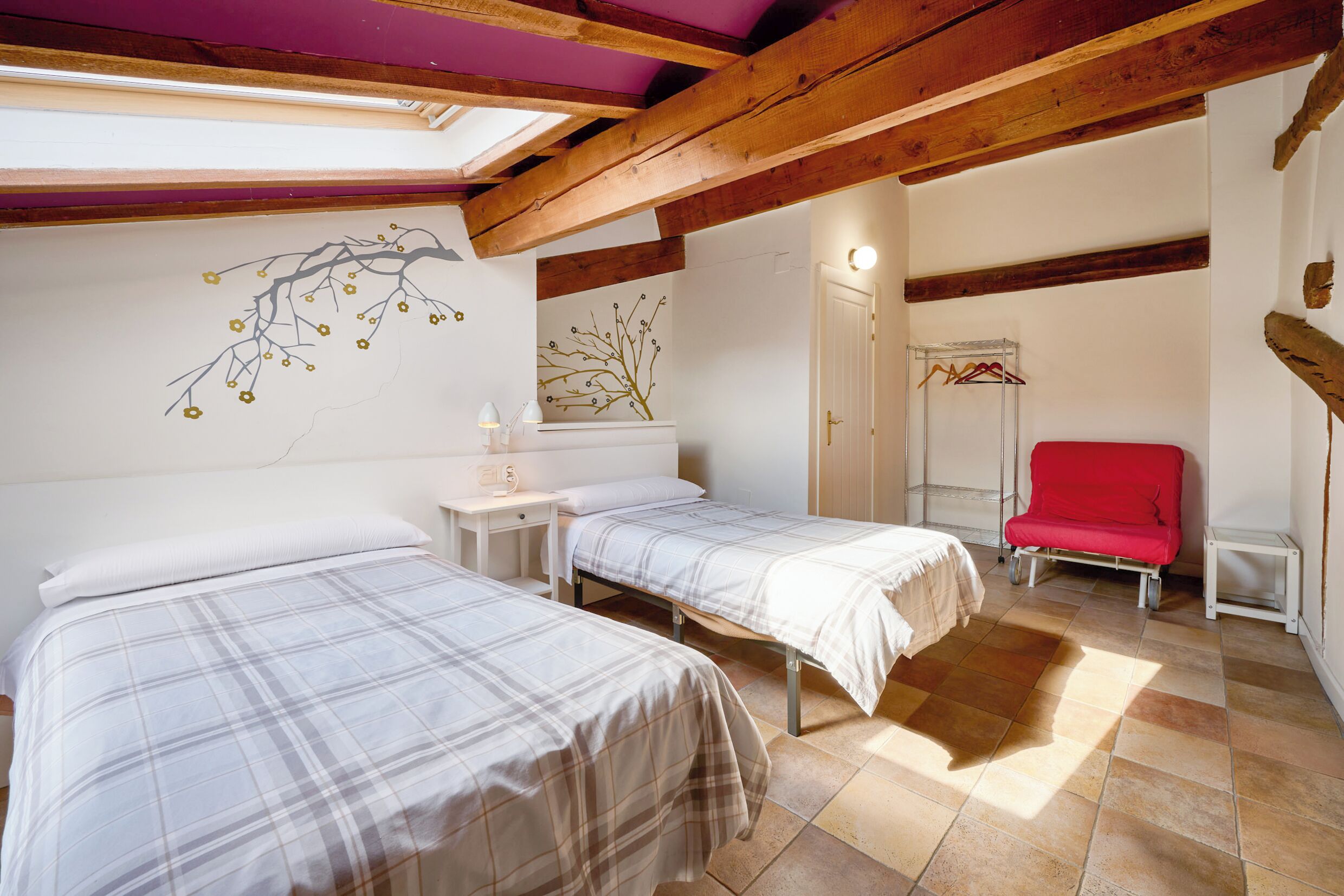 Un dormitorio con dos camas, vigas de madera, un tragaluz, una silla roja y un mural de árbol y flores en la pared.