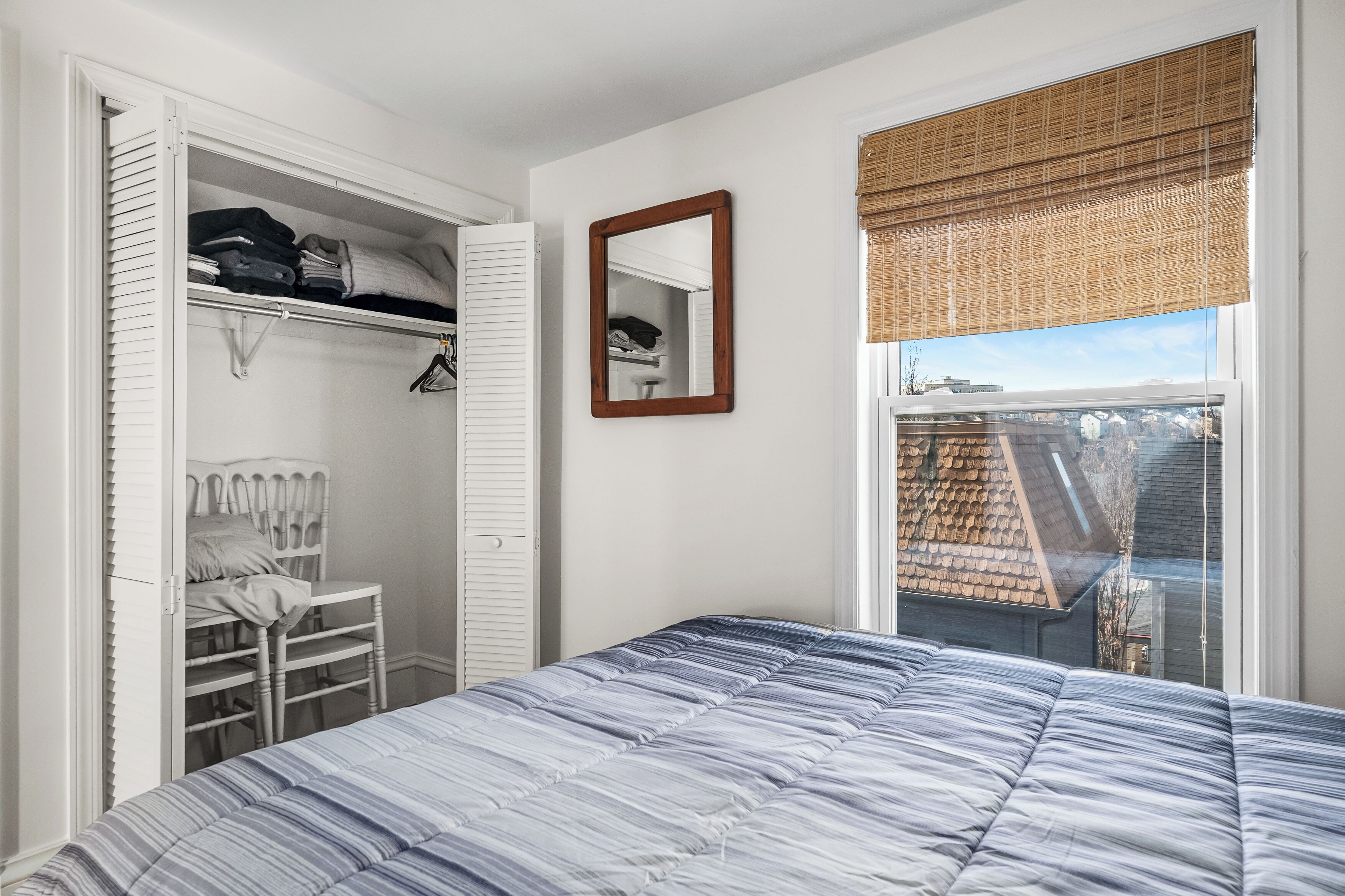 A bedroom with a bed, a wardrobe, a mirror, and a window with a view of buildings.