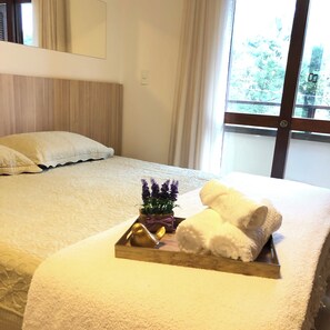 Une chambre d’hôtel avec un lit soigneusement fait, un plateau avec des serviettes et une petite plante, et une fenêtre donnant sur des arbres.