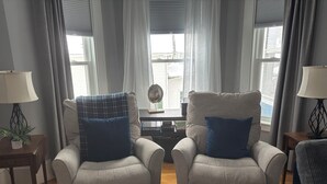 Ein Wohnzimmer mit zwei Sesseln, einem Beistelltisch mit Lampe, einem kleinen Tisch mit Globus und einem Fenster mit Vorhängen.
