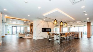 Spaziosa hall d'albergo dal design moderno, con un'area salotto centrale che include un bar, una TV a schermo piatto montata a parete e un'ampia finestra che fa entrare luce naturale.