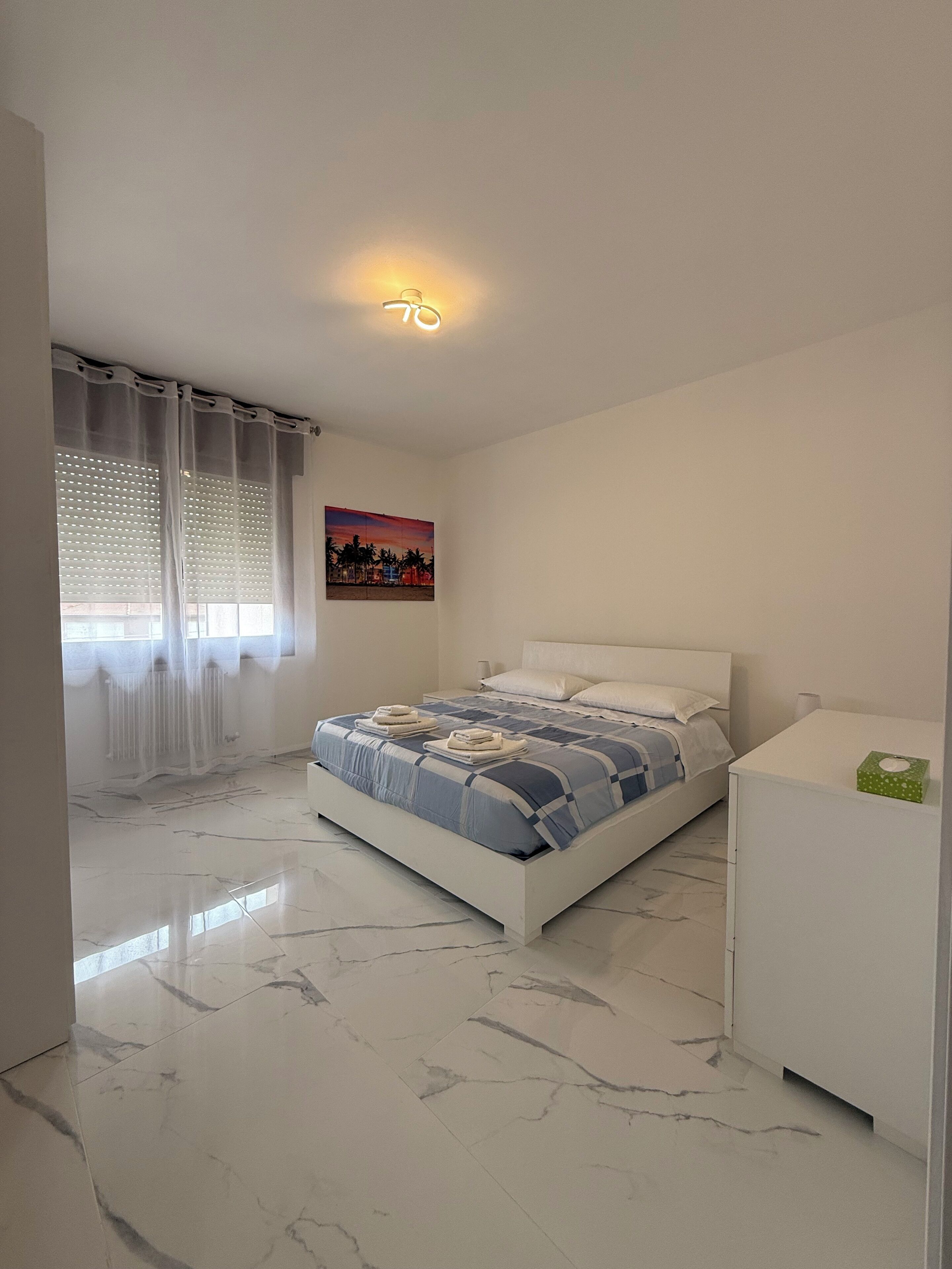 Un dormitorio con cama, mesita de noche, un cuadro y una ventana con cortinas ligeras.