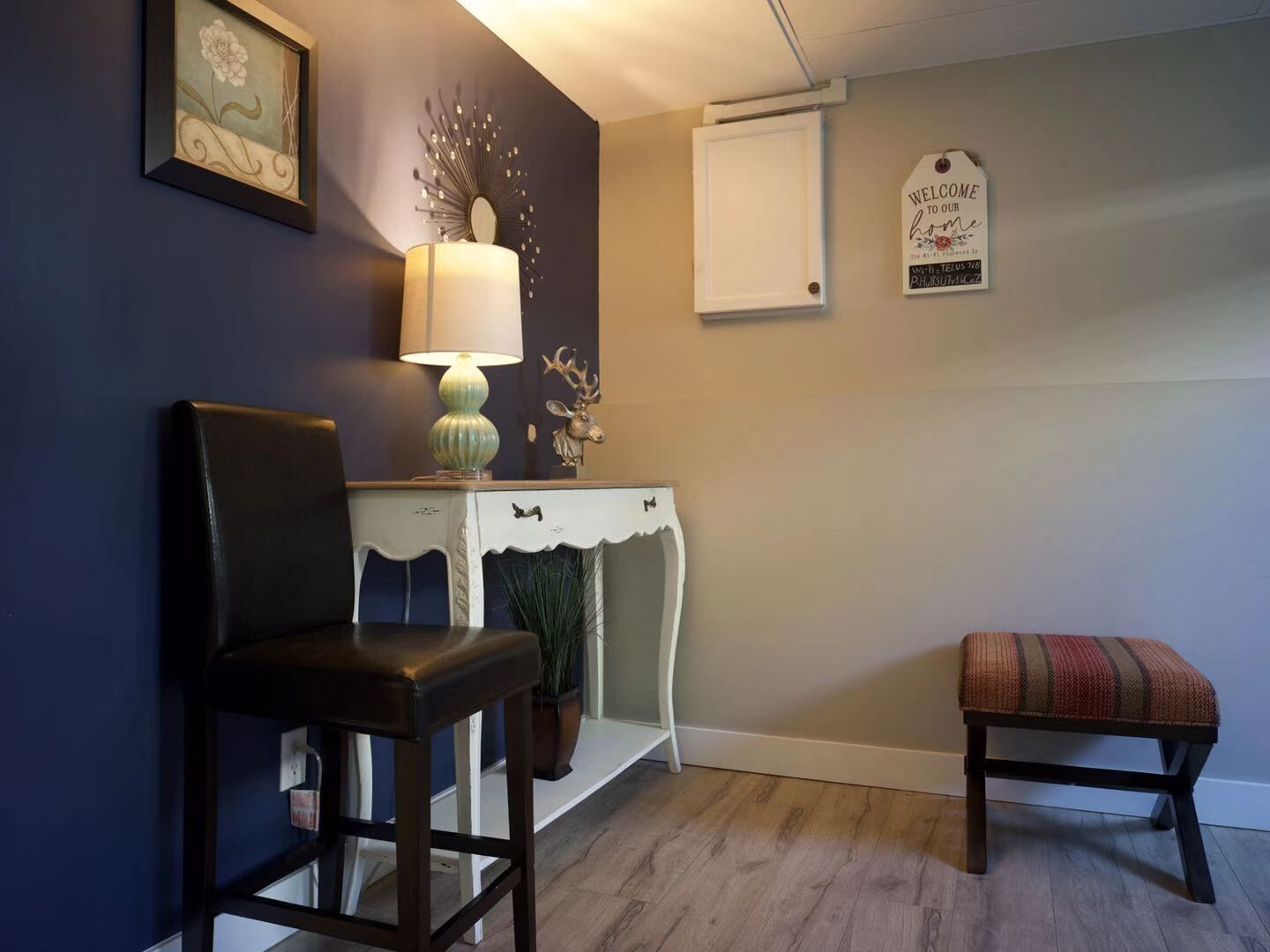 A room with a dark blue wall, a white console table with a lamp, a framed picture, a welcome sign, and a bench.