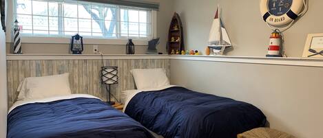 A bedroom with two beds, a window, a shelf with nautical decorations, and a small wooden stool.