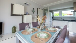 A modern dining area with a table set for two, a vase with flowers, and a kitchen in the background.