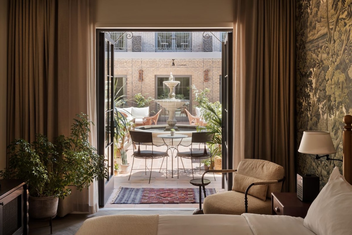 A hotel room with a bed, a desk, a chair, a table with chairs, a small rug, and a view of an outdoor area with a fountain and plants.