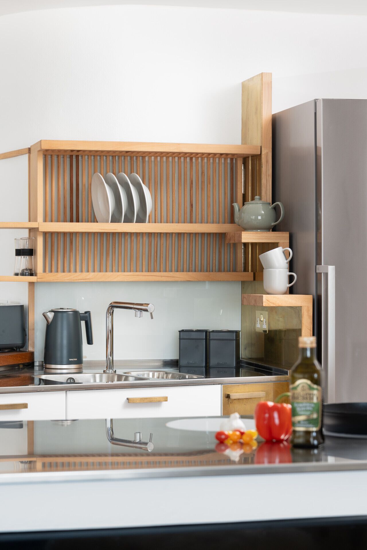 A modern kitchen with wooden shelves, a stainless steel refrigerator, a kettle, and a sink.