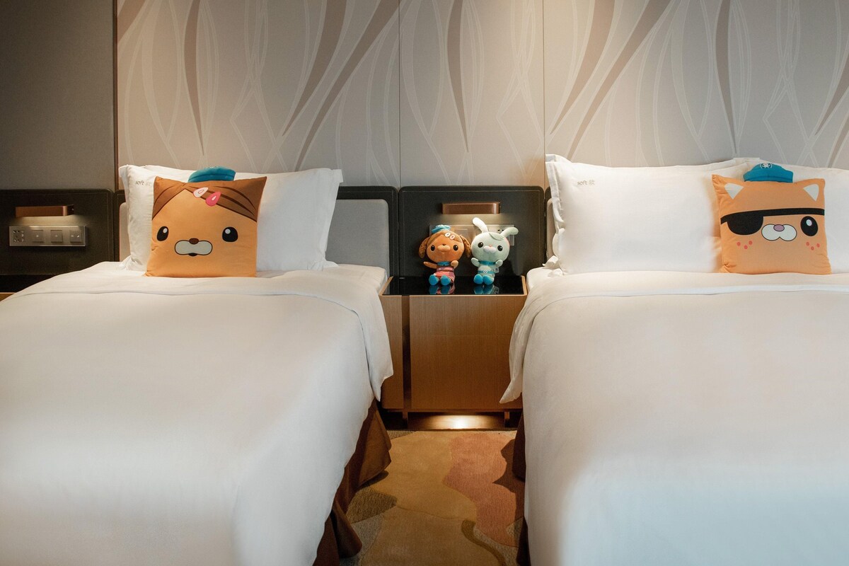 Two beds with plush toys on pillows, a nightstand with a lamp, and a patterned wall.