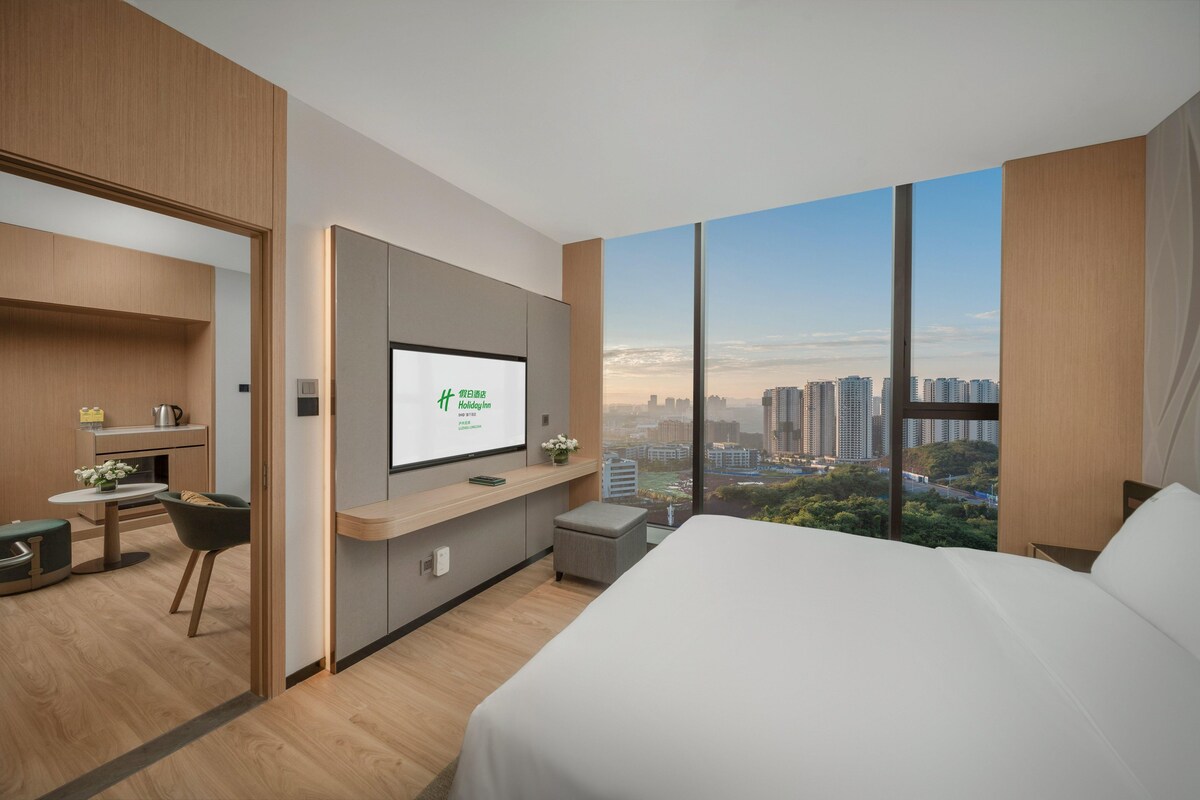 A modern hotel room with a large bed, a desk with a computer, a TV, and a city view.