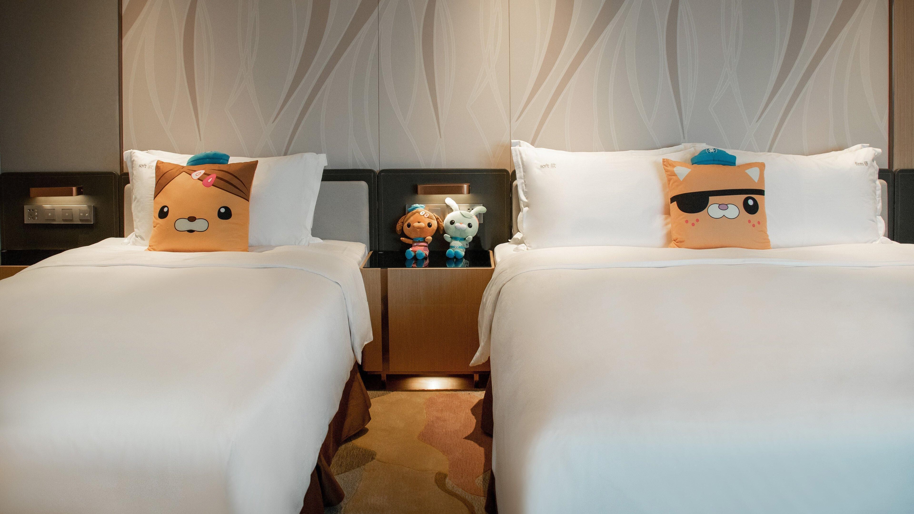 Two beds with plush toys on pillows, a nightstand with a lamp, and a patterned wall.