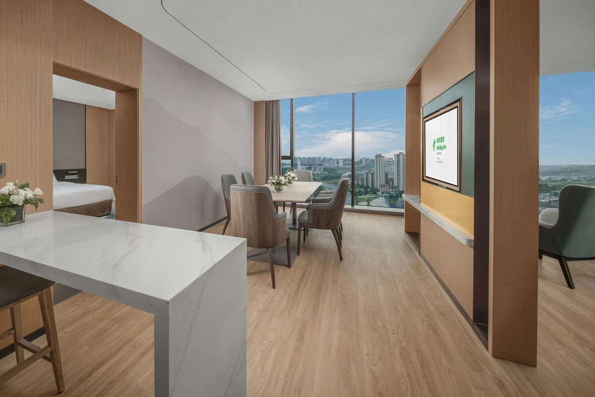 A modern hotel room with a large window, a dining area, and a bed.