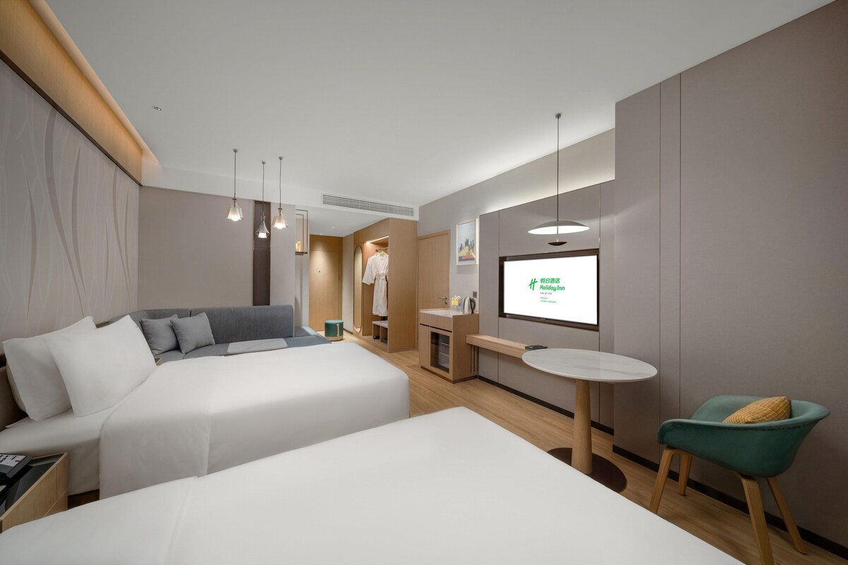 A hotel room with two beds, a desk, a chair, a TV, and a small kitchenette.