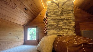 Una habitación de cabaña acogedora con paredes de madera, una pared de piedra con un cráneo de animal montado, una ventana con vistas a árboles y un sofá de cuero marrón con una manta de piel blanca.