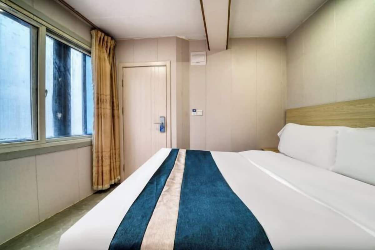 A hotel room with a bed, a window with curtains, a door, and a wall-mounted air conditioning unit.