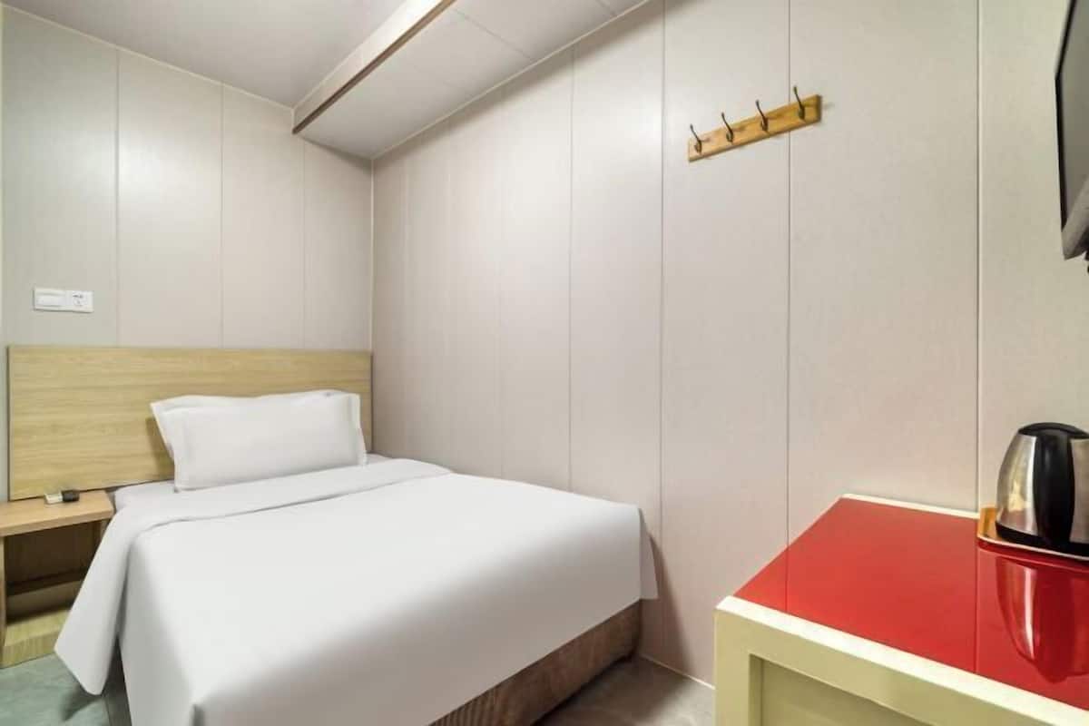 A hotel room with a bed, a wooden headboard, a red table with a kettle, and a wall-mounted hook.