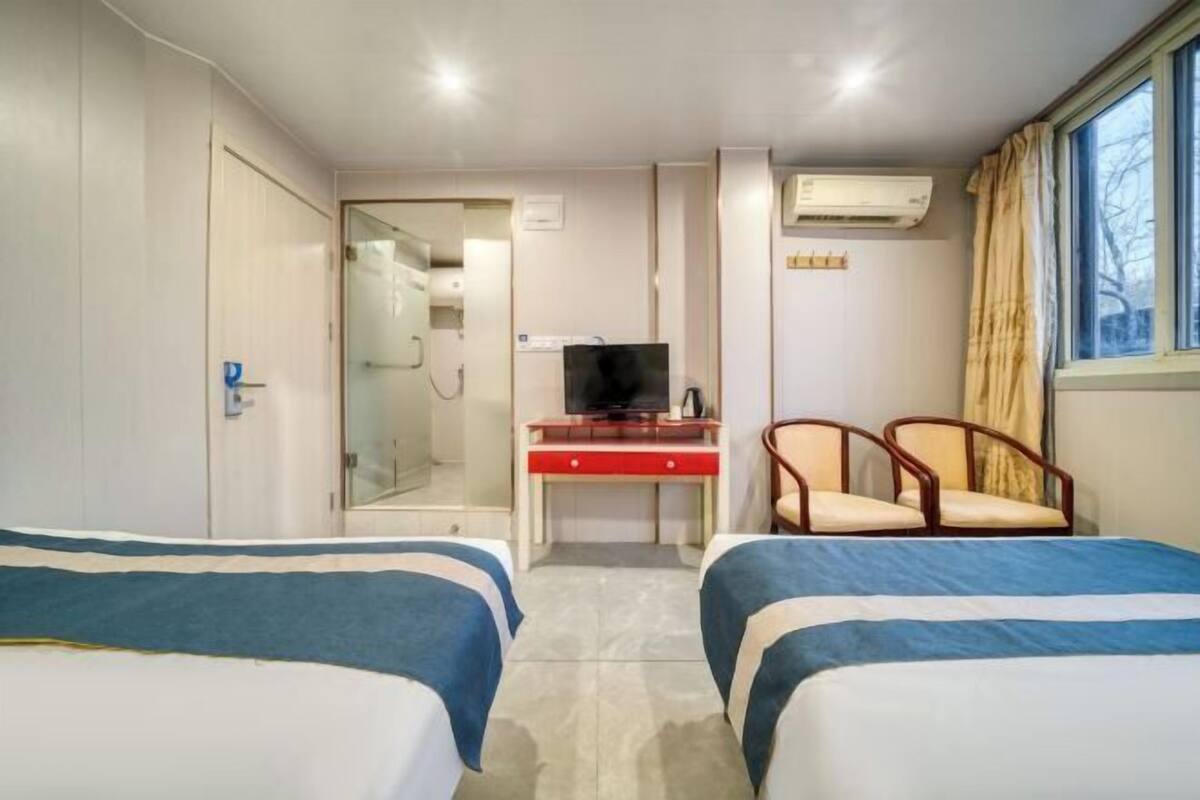 A hotel room with two beds, a desk with a television, a chair, and an air conditioning unit.