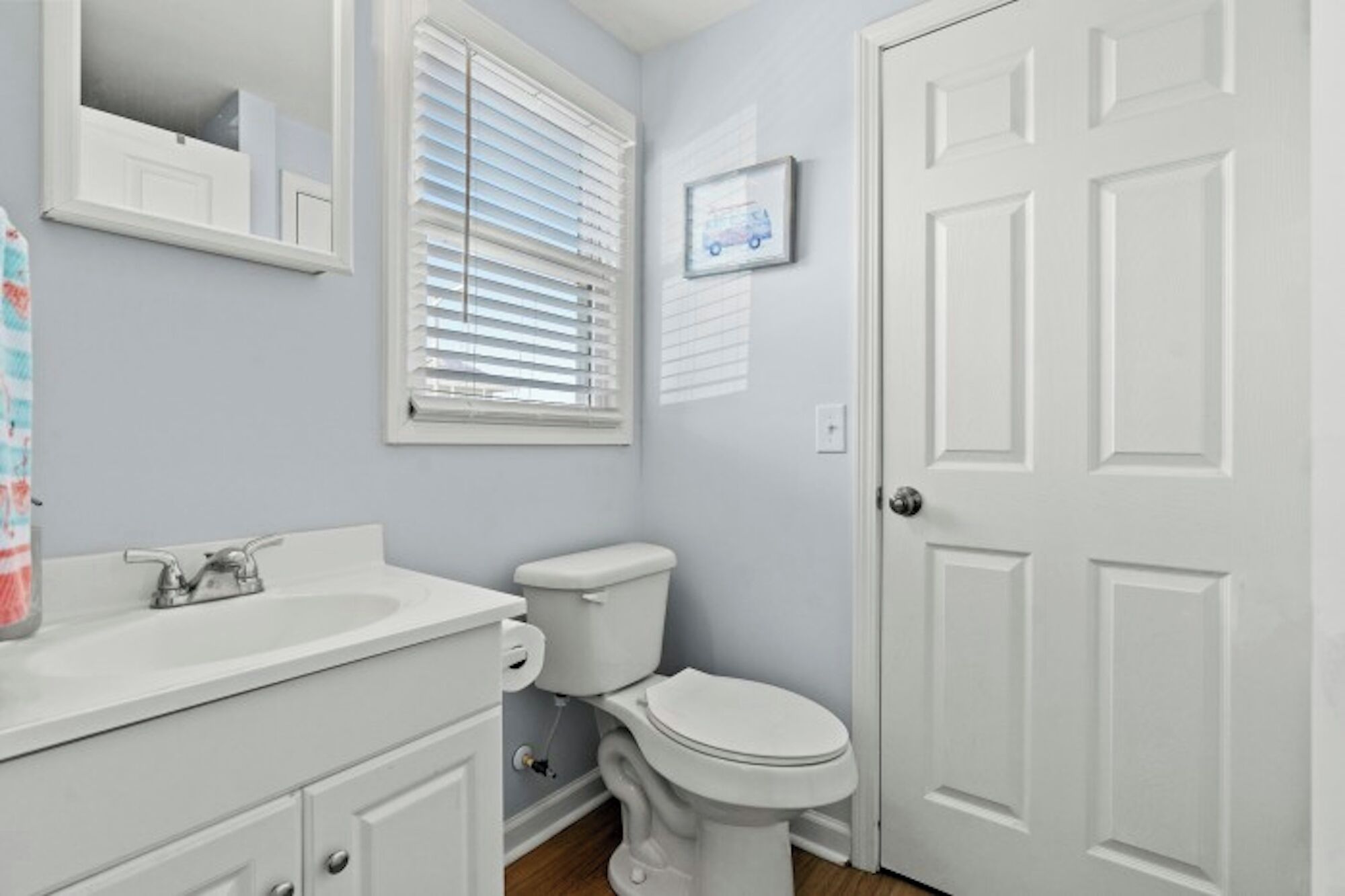 A bathroom with a white sink, toilet, and door. There is a window with blinds and a framed picture on the wall.
