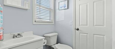 A bathroom with a white sink, toilet, and door. There is a window with blinds and a framed picture on the wall.