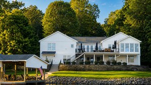 A two-story white house with a dock, a covered deck, and a stone retaining wall. - Lakefront Property! Gentle slope to the water, firepit, water skis, paddleboards (Moneta)