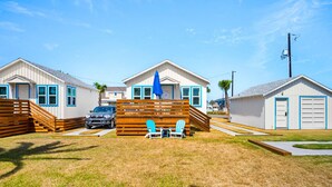 A row of white cabins with blue trim, a wooden deck, and a blue umbrella. - Prime Location in the Heart of Port A – Steps Away from Restaurants, Shops, Parks, and the Beach! (Port Aransas)