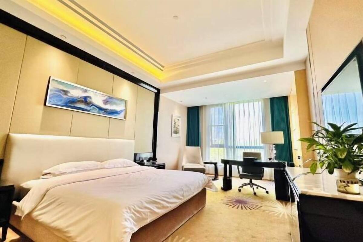 A hotel room with a large bed, a desk, a chair, a desk lamp, a plant, and a painting on the wall.