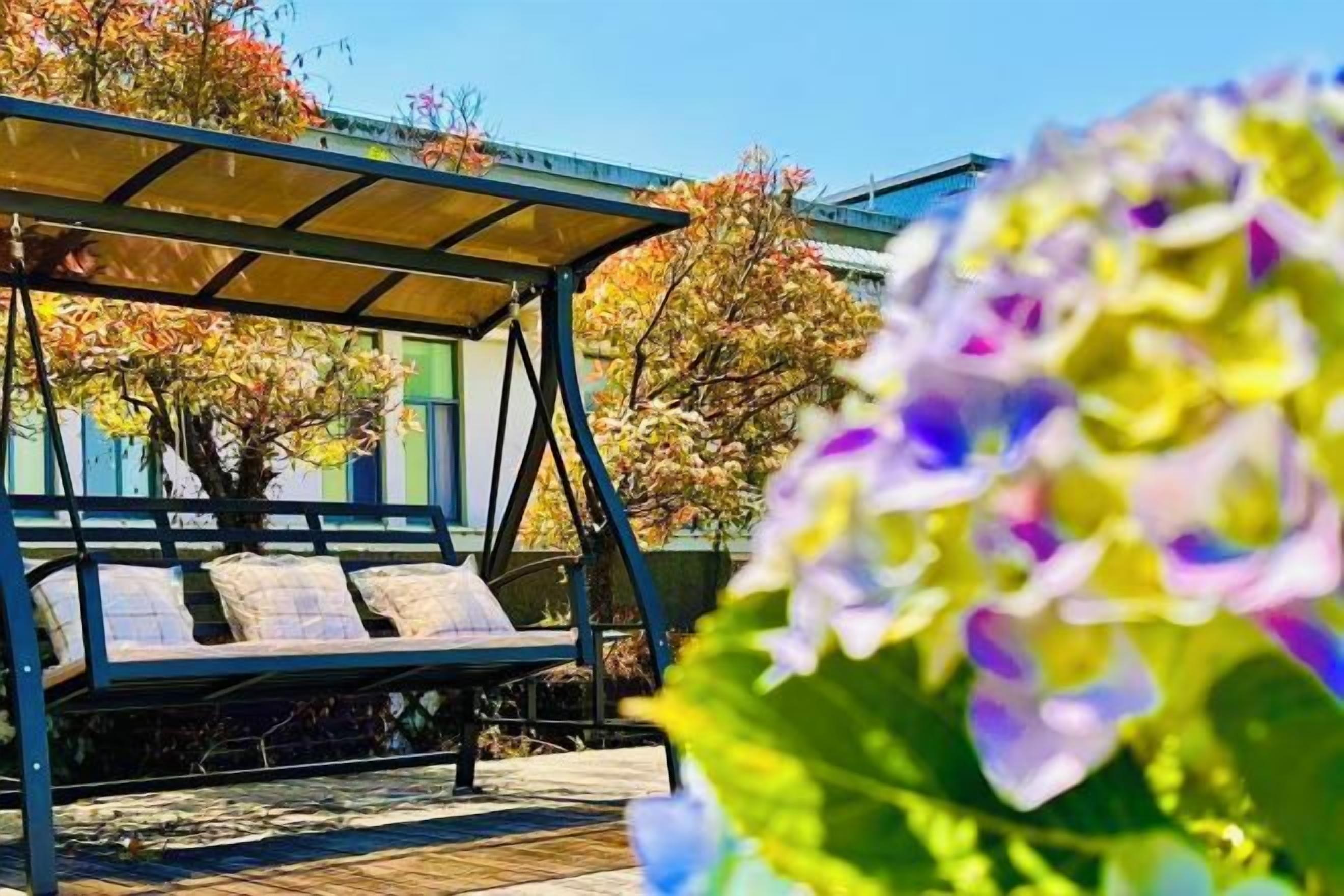 A covered outdoor seating area with a bench, surrounded by trees and flowers.
