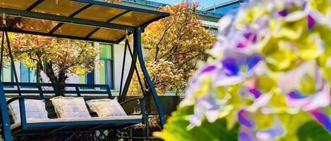 A covered outdoor seating area with a bench, surrounded by trees and flowers.