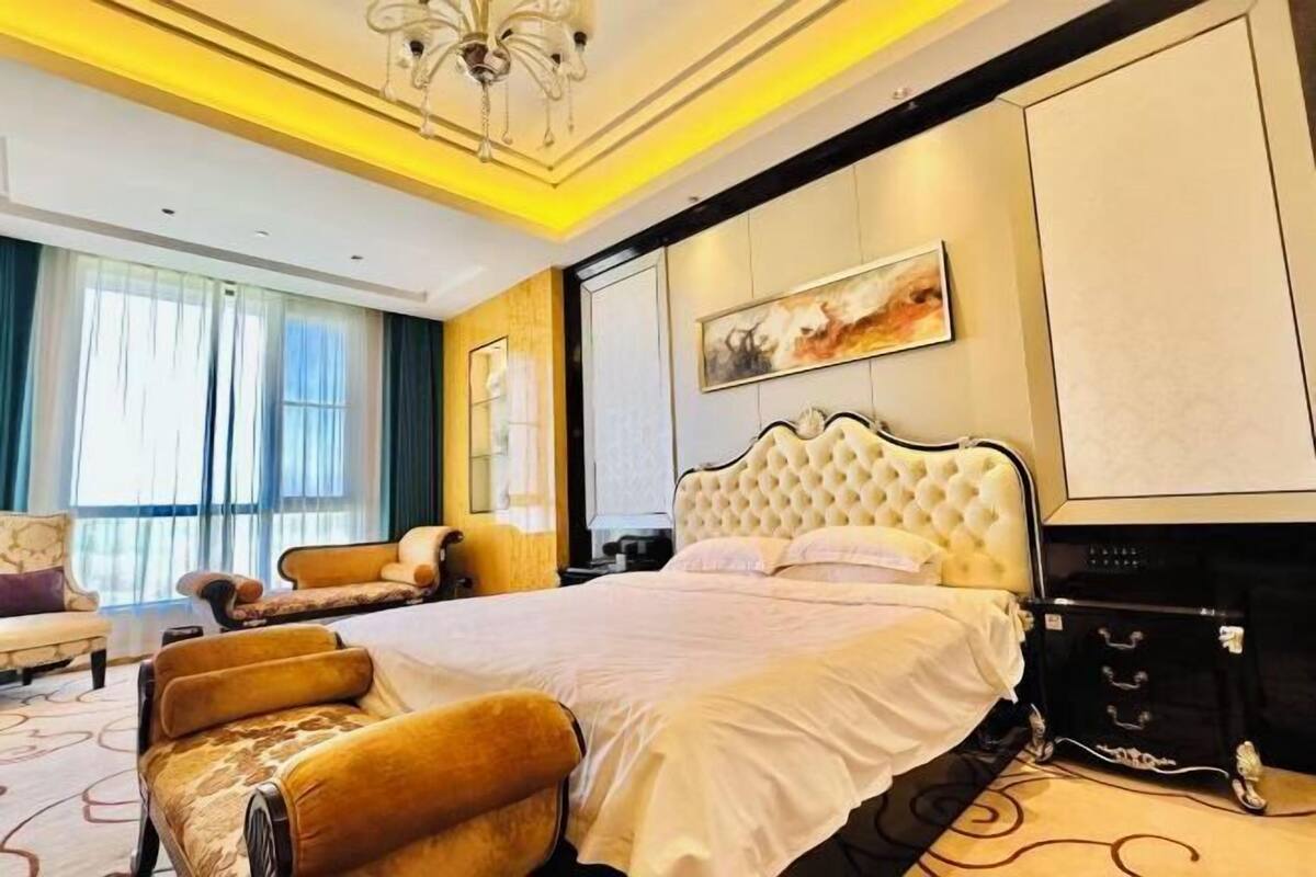 A hotel room with a large bed, a chandelier, a painting on the wall, and a view of the outside.