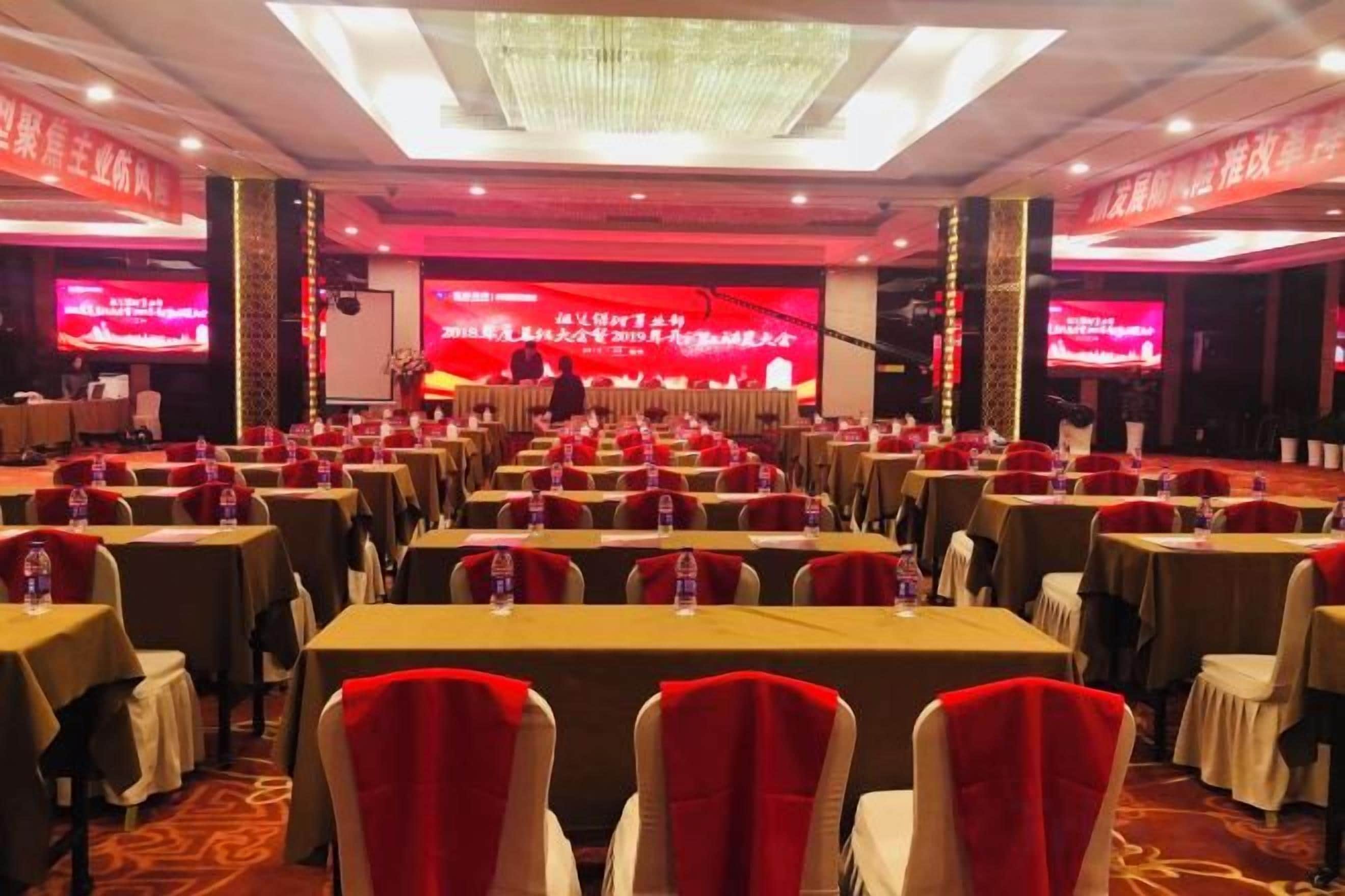 A conference room with rows of tables and chairs, a stage with a large screen, and a chandelier.