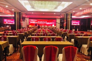 Un salón de conferencias con filas de mesas y sillas, un escenario con una pantalla grande, y una araña.