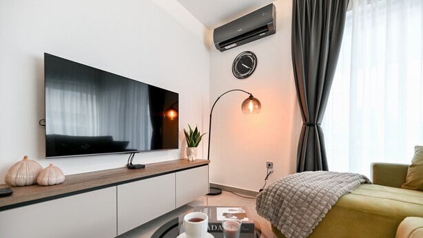 Een moderne woonkamer met een flatscreen televisie, een ronde glazen salontafel met een kopje en kaars, een bank met een plaid, en een vloerlamp.