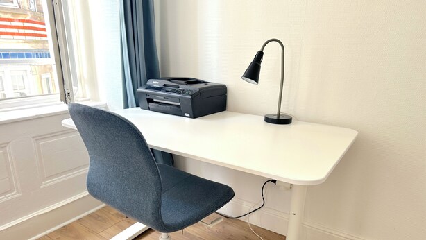 Ein weißer Schreibtisch mit einem schwarzen Drucker, einer schwarzen Schreibtischlampe und einem blauen Bürostuhl.