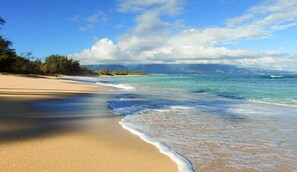 Une plage à l’eau turquoise et limpide, bordée de sable fin, avec des montagnes qui se dessinent à l’horizon sous un ciel partiellement nuageux.