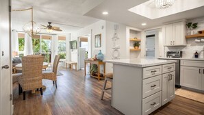 A modern kitchen with white cabinetry, a central island, and a dining area with wicker chairs. - Maui Banyan Retreat | Walk to Kamaole Beach (Kihei)