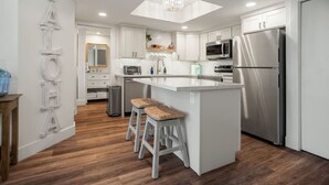 A modern kitchen with white cabinets, stainless steel appliances, a central island with stools, and a skylight. - Maui Banyan Retreat | Walk to Kamaole Beach (Kihei)