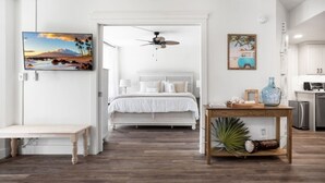 A bedroom with a bed, a ceiling fan, a bedside table, a wooden bench, and a view of the kitchen. - Maui Banyan Retreat | Walk to Kamaole Beach (Kihei)