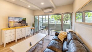 A modern living room with a leather sofa, a white coffee table, a TV on a wooden console, and a sliding glass door leading to an outdoor area. - Walk to Beach! Pool, Hot Tub + AC Studio (Kihei)