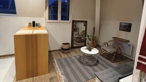 Un espace de vie compact comprenant une estrade en bois, une table ronde, une chaise en osier, un tapis à motifs et une fenêtre donnant sur un immeuble.