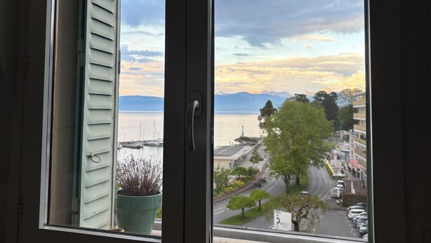 Une vue depuis une fenêtre, donnant sur une étendue d’eau, un arbre et des bâtiments au loin.