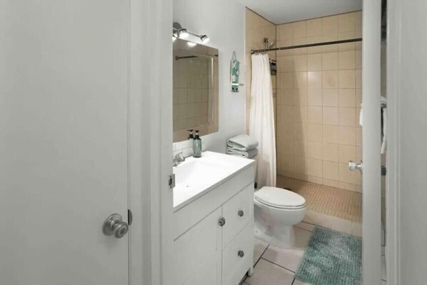 A bathroom with a white sink, mirror, and toilet, a shower with a beige tiled wall, and a green rug on the floor. - Kihei Escape | Walk to Beach, Pool & Hot Tub (Kihei)