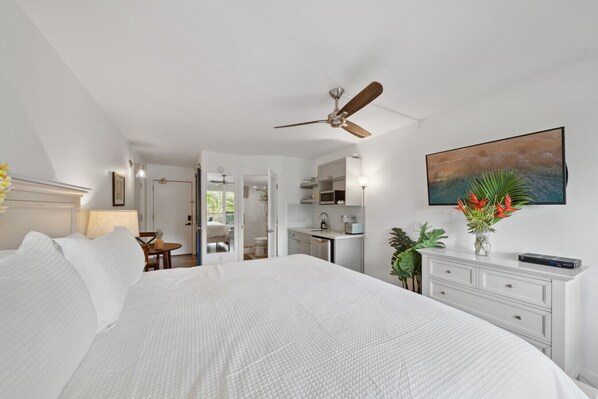 A bedroom with a large bed, a ceiling fan, a bathroom with a shower, and a kitchen area. - Steps to Beach | Maui Banyan King Studio (Kihei)